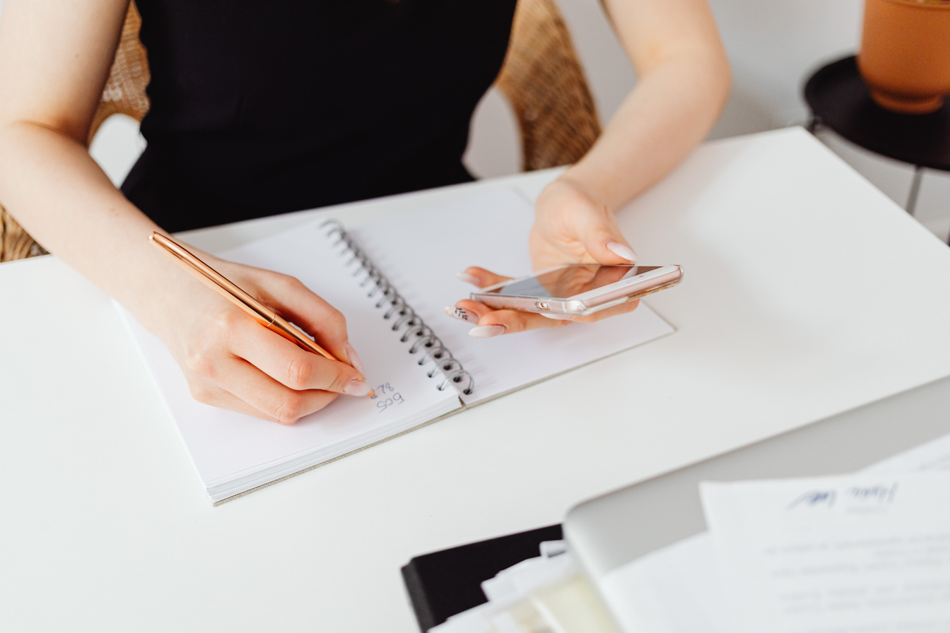 A Person Writing on a Notebook while Holding a Cell Phone